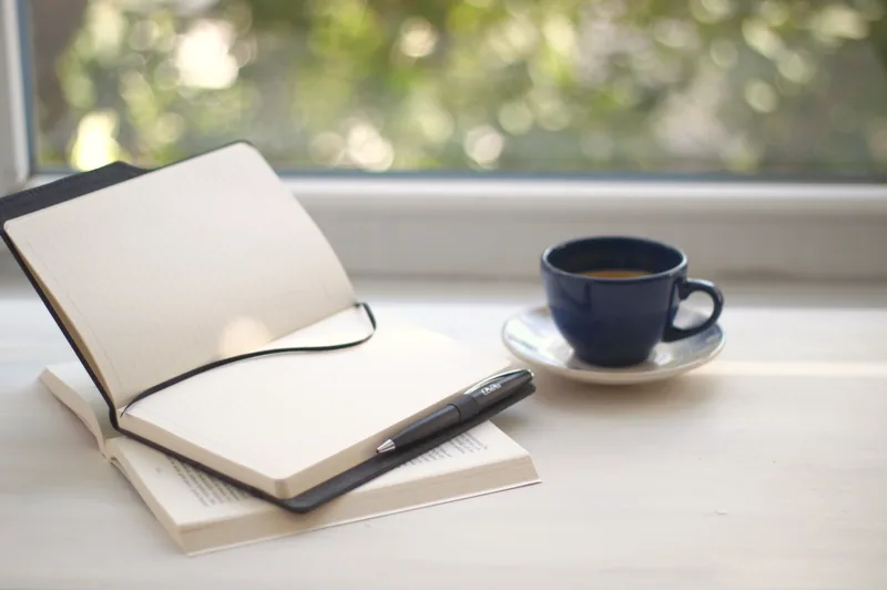 An open notebook and a cup of coffee on a desk by a window